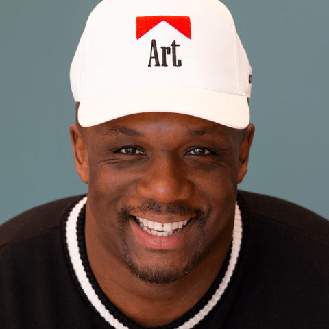 Harold is a Black man. He wears a white cap with "Art" in place of the word "Marlboro" in a Marlboro cigarette logo.