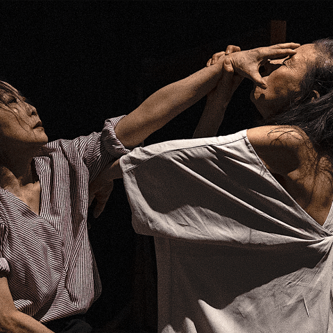 An Asian woman holds her hand over the face of another Asian woman, whose back is exposed in a loose shirt.