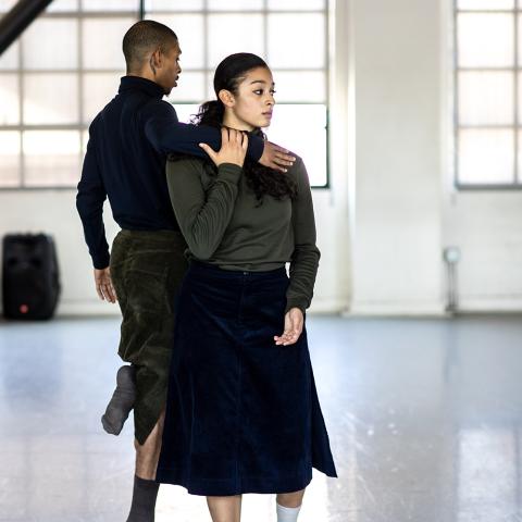 In an empty, windowed space, two folks of color dance in long black skirts.