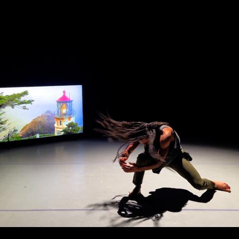 In front of a screen with a lighthouse projection, a Black man throws his braids to the right as he bends to one knee.