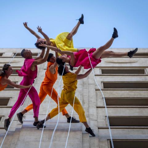 On the side of a building, looking up, six performers are strung to the building in bright, warm tones. They appear to hold two of the dancers up off the building, defying gravity.