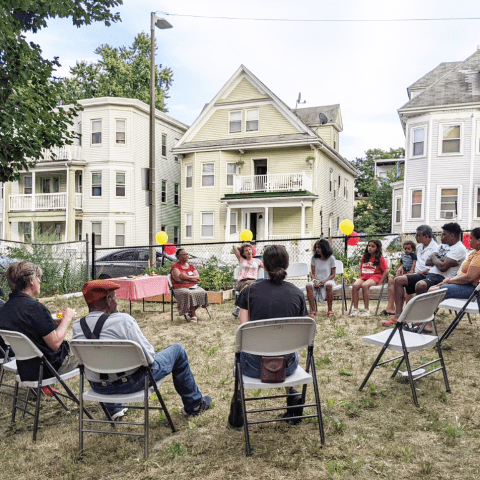 a group of people sitting in a circle in folding chairs in a neighborhood park. Balloons decorate a few of the chairs.