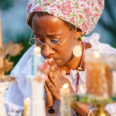 A brown woman prays to her hands, with candles lit in the foreground. 