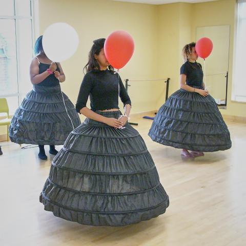 Four women in black dresses each hold a single red or white balloon in front of their faces in a dance studio.