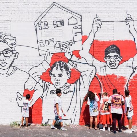 Six kids hang out by a mural of three kids of varying ethnic backgrounds hold up their homes as picket signs.