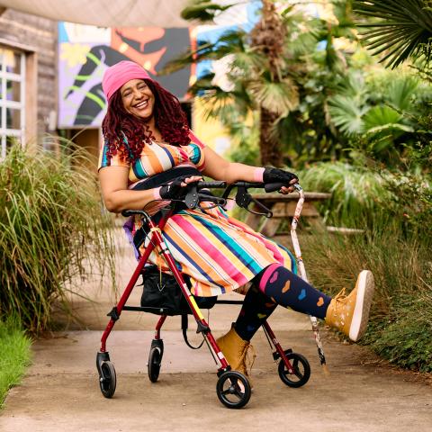 Claudia laughs while sitting back in a rollator, their right hand holding a cane and their left leg kicked out casually. They wear a beanie hat, red braids, bike gloves layered over arthritis gloves, a rainbow striped dress, rainbow heart leggings, and sneakers. Claudia is in an outdoor courtyard with a paved walkway, tropical foliage, and overhead shade sail covers. 