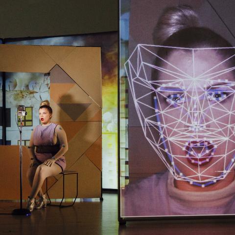 A woman on a stage sits next to a live video of herself that is close on her face and has lines mapped to parts of her face.