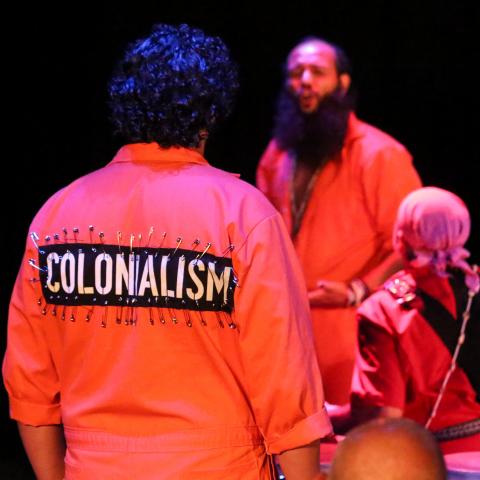 A man in an orange jumpsuit has pinned the word "Colonialism" to the back of it.