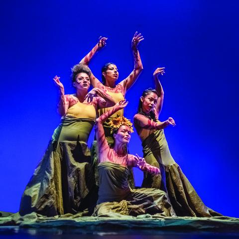 Four women in a diamond formation dance in front of a blue backdrop.
