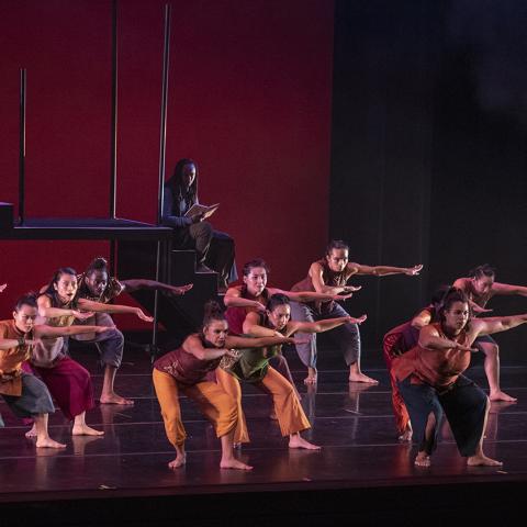 On a stage with a red backdrop, a group of women lean to their right and hold their left arm level with their shoulder.