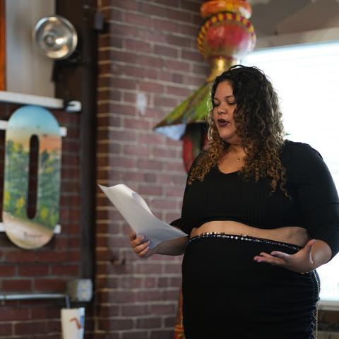 a brown woman with curls reads from a sheet of paper in a restaurant.