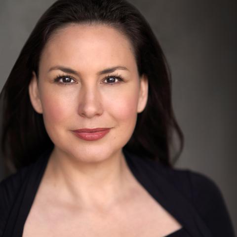 DeLanna is an Indigenous woman with long brown hair and black eyes. She is wearing a black shirt with short sleeves. She stand in front of a charcoal grey background. 