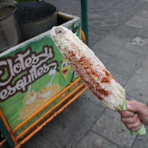 A woman holds an elote in front of a cart that also says, "Elotes."