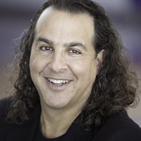 A man with long curly hair and sporting a black suit jacket smiles.