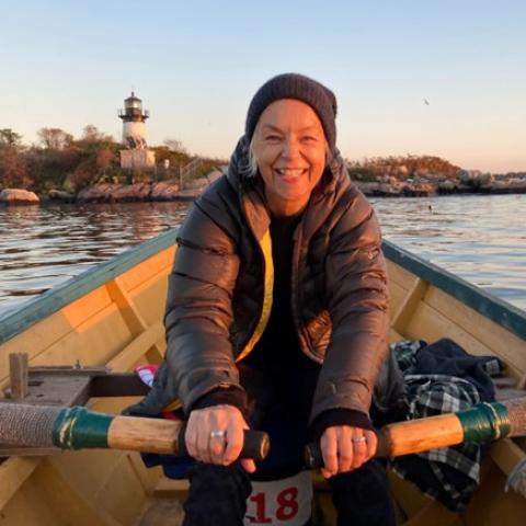 Erica paddles a boat. She's a white lady with white hair and a parka. A lighthouse is behind her.