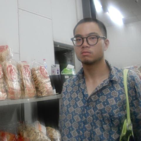 Che has short hair. He has big black glasses and wears a checkered shirt. He is an Asian man. He poses next to bread in plastic bags.