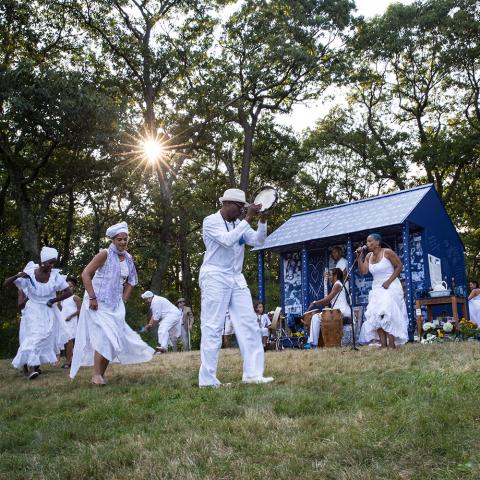 Ife stands in front of a cabin that is wrapped in fabrics dyed in indigo. A crowd, dressed all white, dance around her.