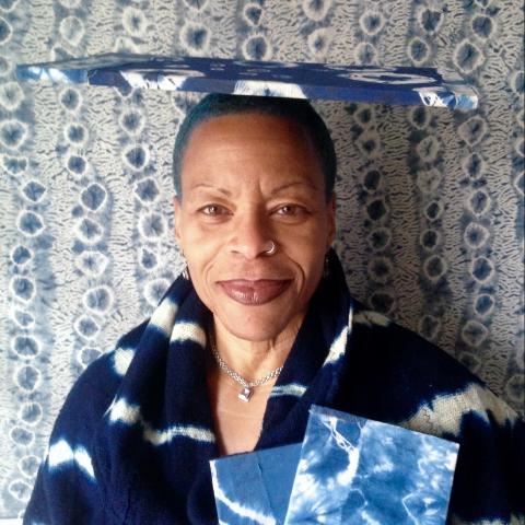 Ife wears blue and poses with a blue package on her head in front of a wall with a blue floral wallpaper.