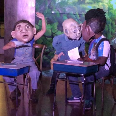 Wearing over-sized masks, three performers sit behind desks.