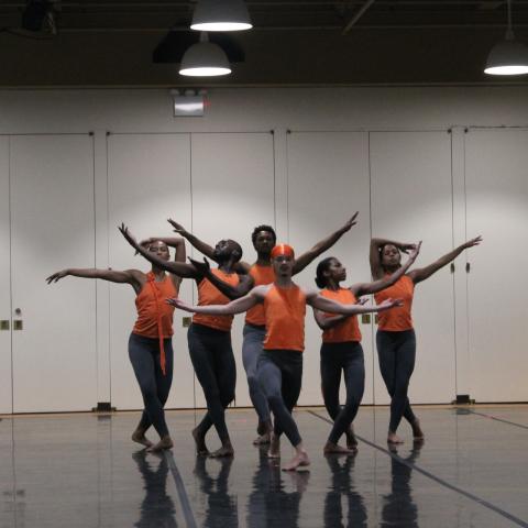 Black, male dancers in a studio space. They wear fitted, orange tank tops and grey pants.