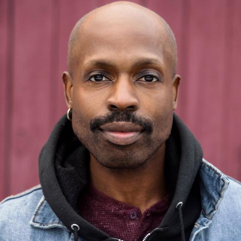 Jarvis is a bald, black man with a moustache. He wears a black hoodie under a jean jacket.