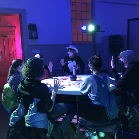 A group of 6 people sit around a table in a semi-circle surrounding a person with brown skin, a dark shirt, and a white cowboy hat. The table has a dark orb sitting in the center, and a variety of papers around it. The room is lit in dark blue lighting with a pink glow coming from a room behind. The edge of a projection screen showing numbers and a starscape can be seen to the right.