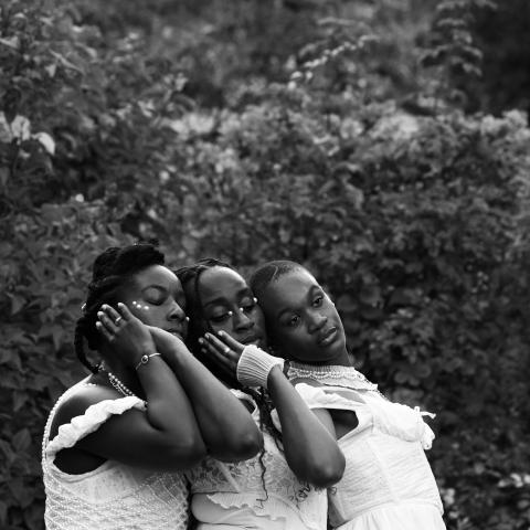 In a garden, three Black women caress each other. They're all in white gowns.