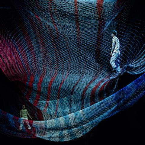 On colorful, transparent netting, two dancers walk towards each other.