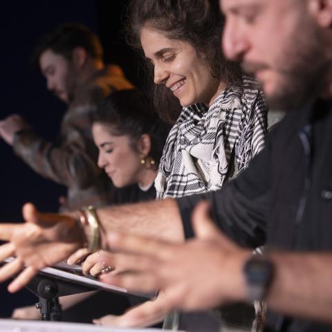 A close up on four actors at music stands, against a black background, gesturing expressively with their hands as they read from scripts. 
