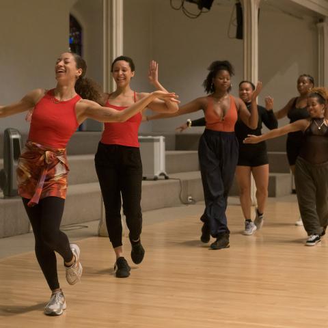 Six women stand in a row with their arms out and their feet in motion, walking toward the camera.