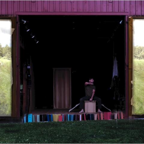 Outside, a man performs in a barn, where the doors have projections of fields on them.