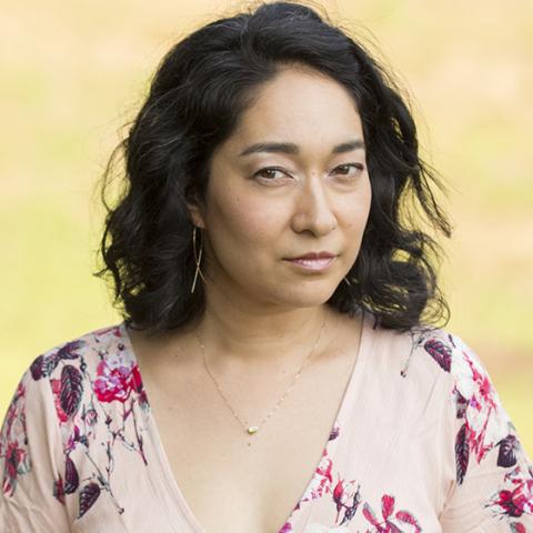 An image of Meena, a South Asian and East Asian woman, with black wavy shoulder-length hair, wearing gold earrings, gold necklace and a v-neck floral pink dress. The yellow and green background is blurred. 