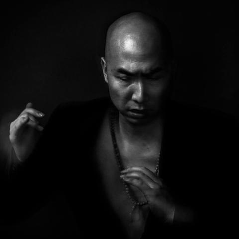 A black and white photo of a bald, Asian man who wears an open-chest, soft shirt and looks down.