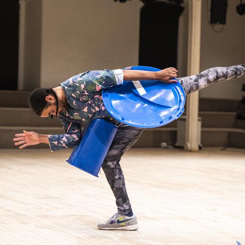 A Black man, with braids, wears a floral body suit and holds or wears blue, pastic cans and lids. He leans on one leg.