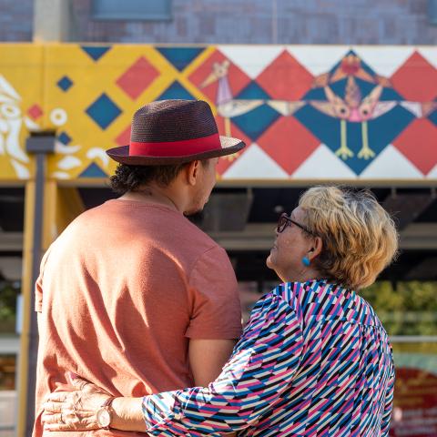 From behind them - a man, in a fedora, is held by an older woman with blonde highlights. They look up at a mural along the top of a building.