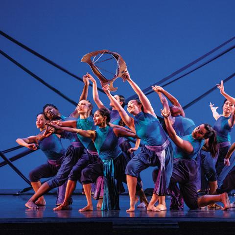 On a stage, a troupe of dancers wear blue and purple in front of a blue backdrop.