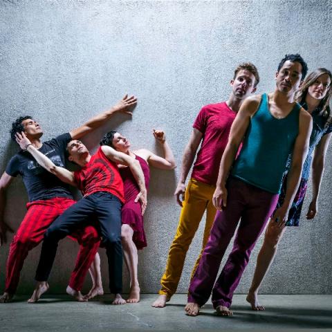 Three dancers wearing bright colors lean against a wall while three more lean to the left in the foreground.