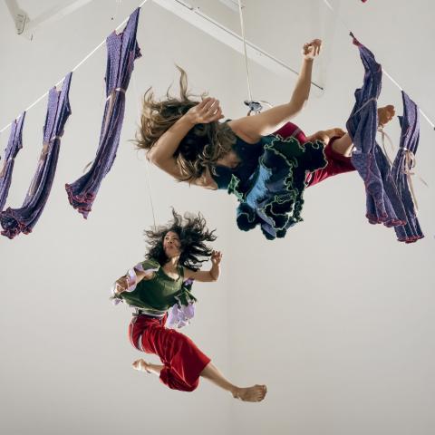 Two women dance between hanging laundry in a white walled studio space.