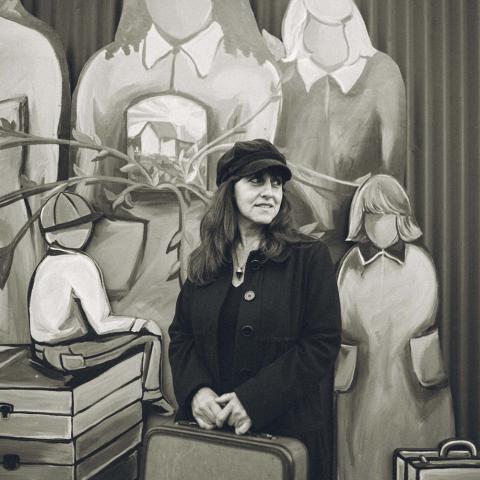 Nora poses in front of a painting of faceless figures with suitcases while holding a suitcase.