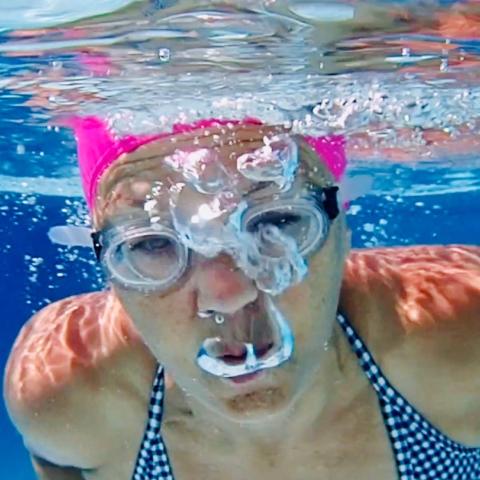 Underwater, a white woman swims in a pink swimming cap.