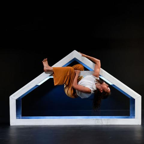On a stage, a woman hangs from a frame that is shaped like a short house.
