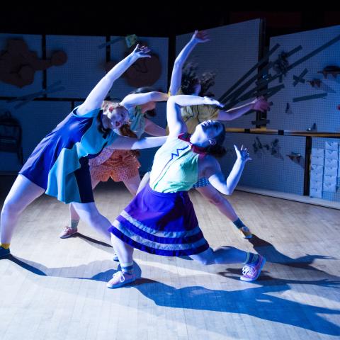 In front of floral backdrops, three dancers lean back in multi-colored outfits.