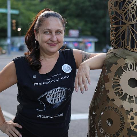 Ruth has light skin and long brown hair with red highlights. She leans on a pole with one a sculpture of gold gears wrapped around it.