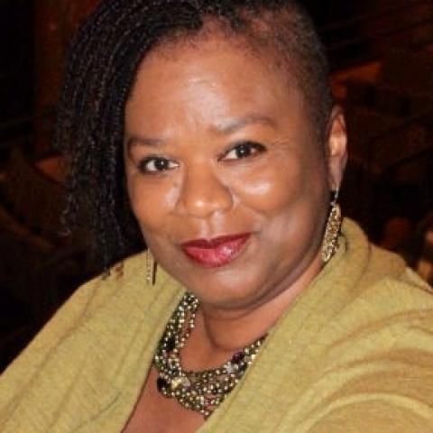 A woman with braids on one side and beaded earrings and necklace, wearing a yellow sweater