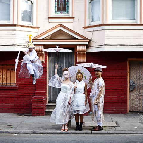 Outside a San Franciscan home, a person dances on a stoop while the others wear white lace and hold white umbrellas.