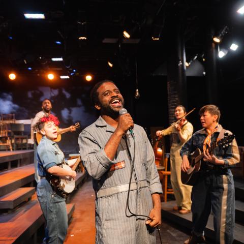 Five folks perform. A Black man sings at the center with the rest of the band behind him. There's a stage behind them to the left and steps in both directions.