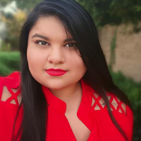 Very long, blue-black hair, light brown skin, red lipstick wearing a red blouse in front of a blurred outdoor background.