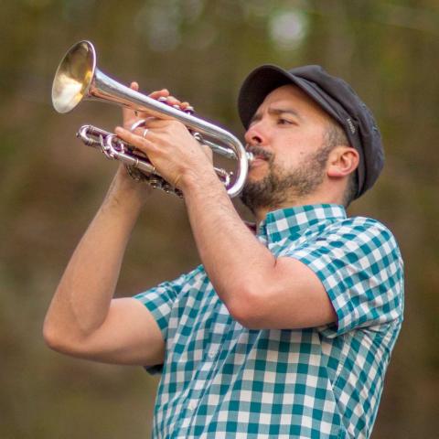 A man in a plaid shirt playing the trumpet outside with trees in the background