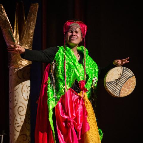 Terry wears a costume with bright green and pink and yellow fabric. The pink fabric is wrapped around her head and she holds wooden sculptures.