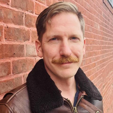 a white man with short hair and a mustache stands in front of a brick wall